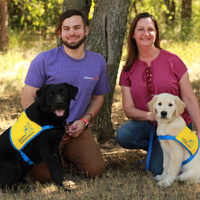 A man sits in a field with a black Labrador sitting in front of him, while a woman sits with a yellow Labrador puppy sitting in front of her.