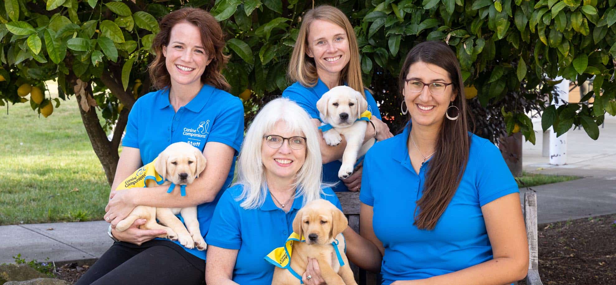 Four smiling women in canine companions polos holding puppies in yellow puppy capes