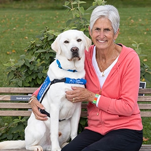 A smiling woman sitting next to a yellow lab service dog in a blue canine companions vest