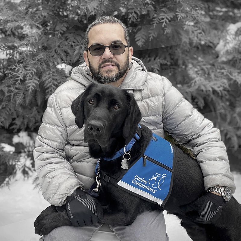 A man in sunglasses with a black lab in a blue service vest laying across his lap