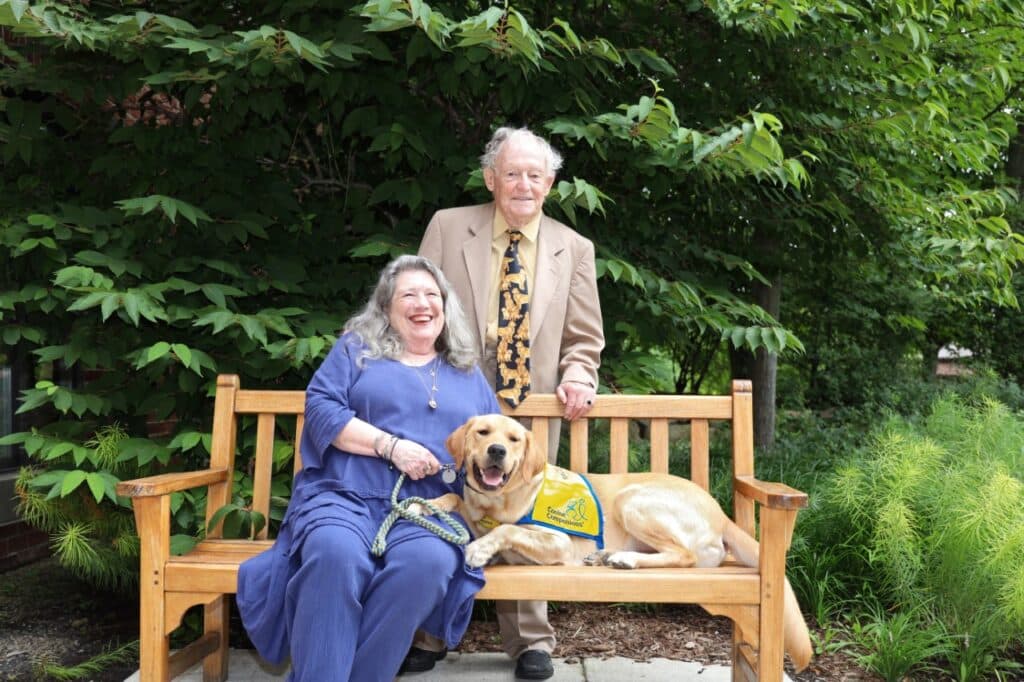 A woman sits on a bench with a yellow lab wearing a blue and yellow vest while her husband stands behind the bench and smiles.