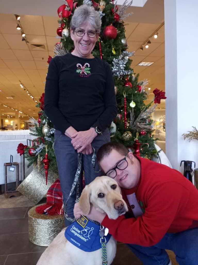 A young man hugs his service dog with his mother standing behind