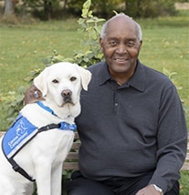 A smiling man sitting next to a yellow lab wearing a blue canine companions vest