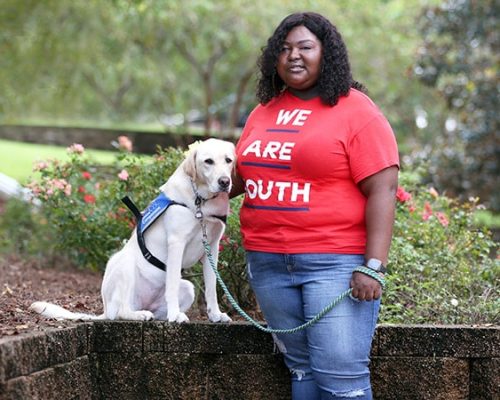 juniorambassadorboard2 A Smiling young African American standing next to a yellow lab wearing a blue service vest