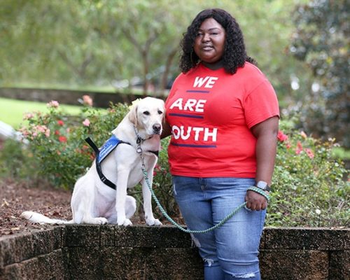 A Smiling young African American standing next to a yellow lab wearing a blue service vest