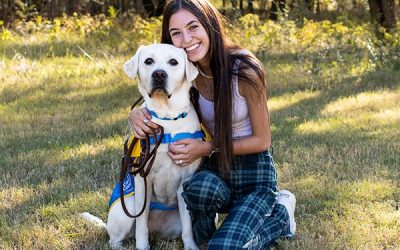 A young girl kneeling on the grass hugging a yellow lab wearing a yellow puppy vest