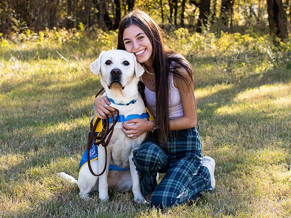 A young girl kneeling on the grass hugging a yellow lab wearing a yellow puppy vest