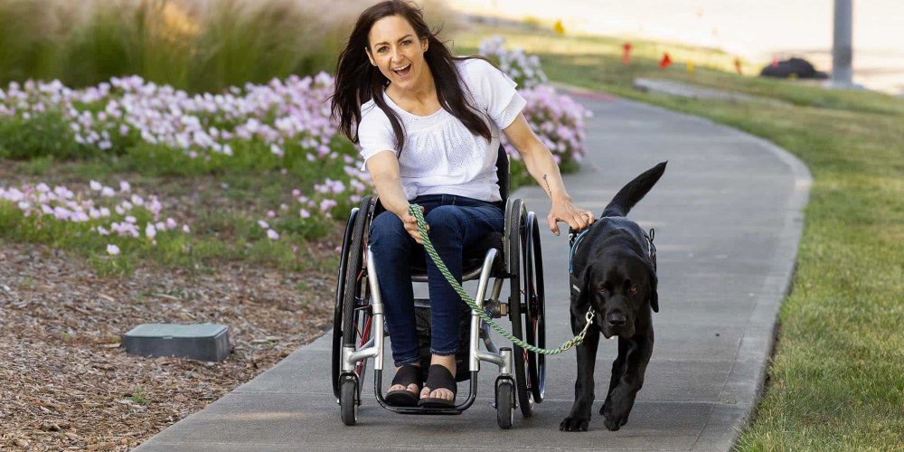 mackenzie-pull a woman in a wheelchair being pulled along a park path by her black lab service dog