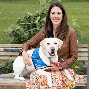 A smiling woman sitting next to a yellow lab service dog in a blue canine companions vest