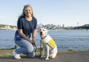 a smiling young woman with a yellow lab in a yellow puppy vest on the Seattle bayfront