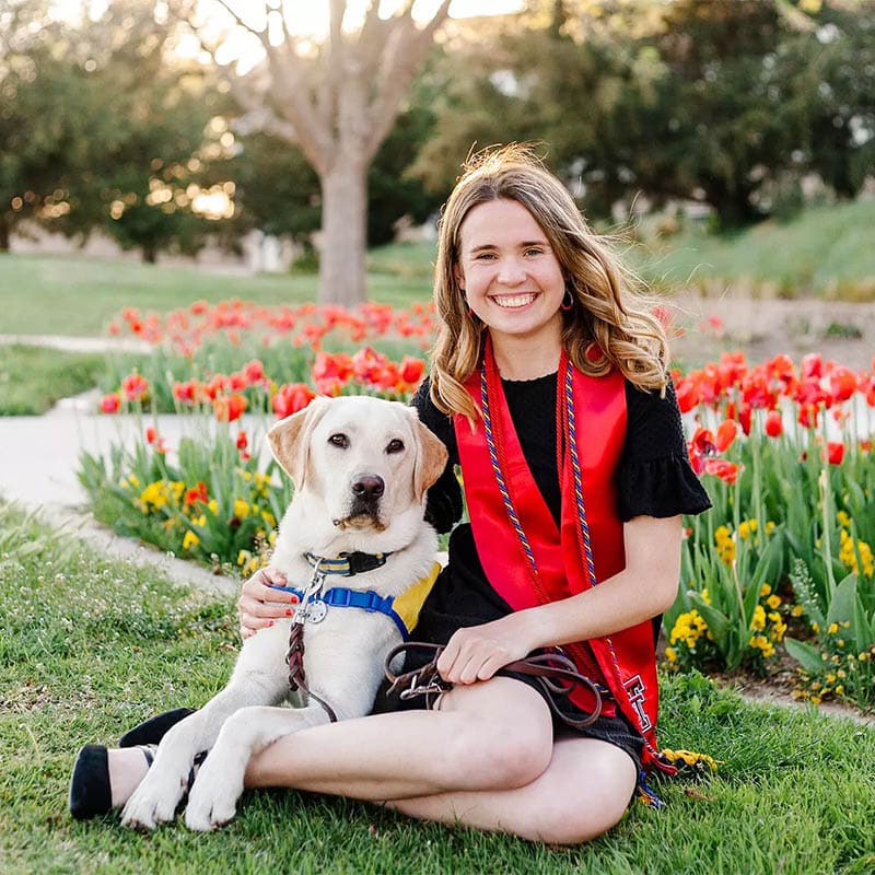 a smiling girl sitting next to a yellow lab in a yellow puppy vest