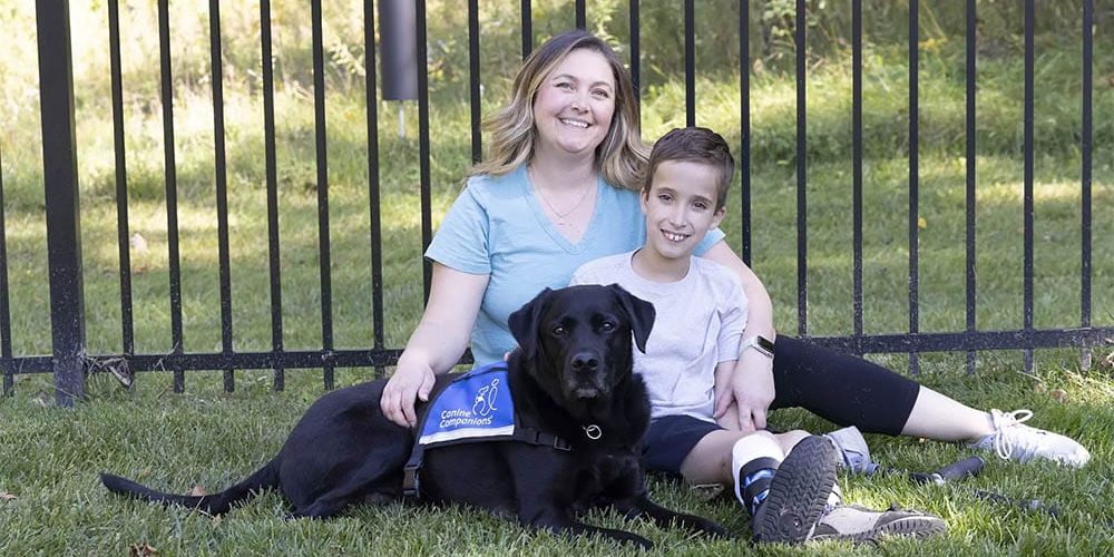 A young boy sits on the grass with his mother and his black lab service dog wearing a blue vest