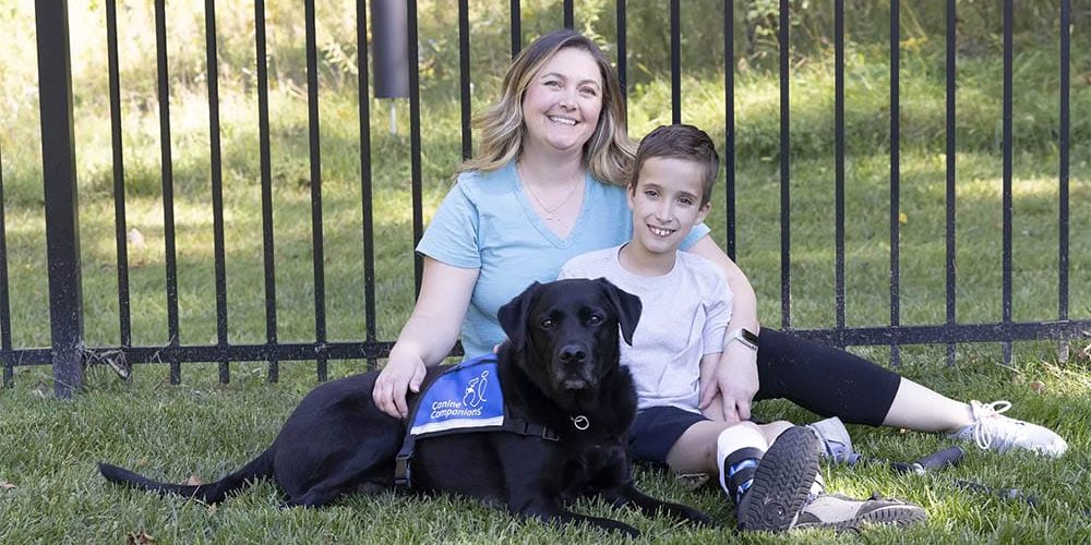 stryderandfonz A young boy sits on the grass with his mother and his black lab service dog wearing a blue vest