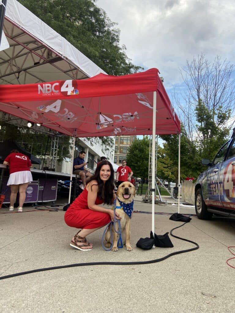 A woman kneels next to a yellow lab wearing an American flag bandana. The two are under a red awning that reads ‘NBC4.’