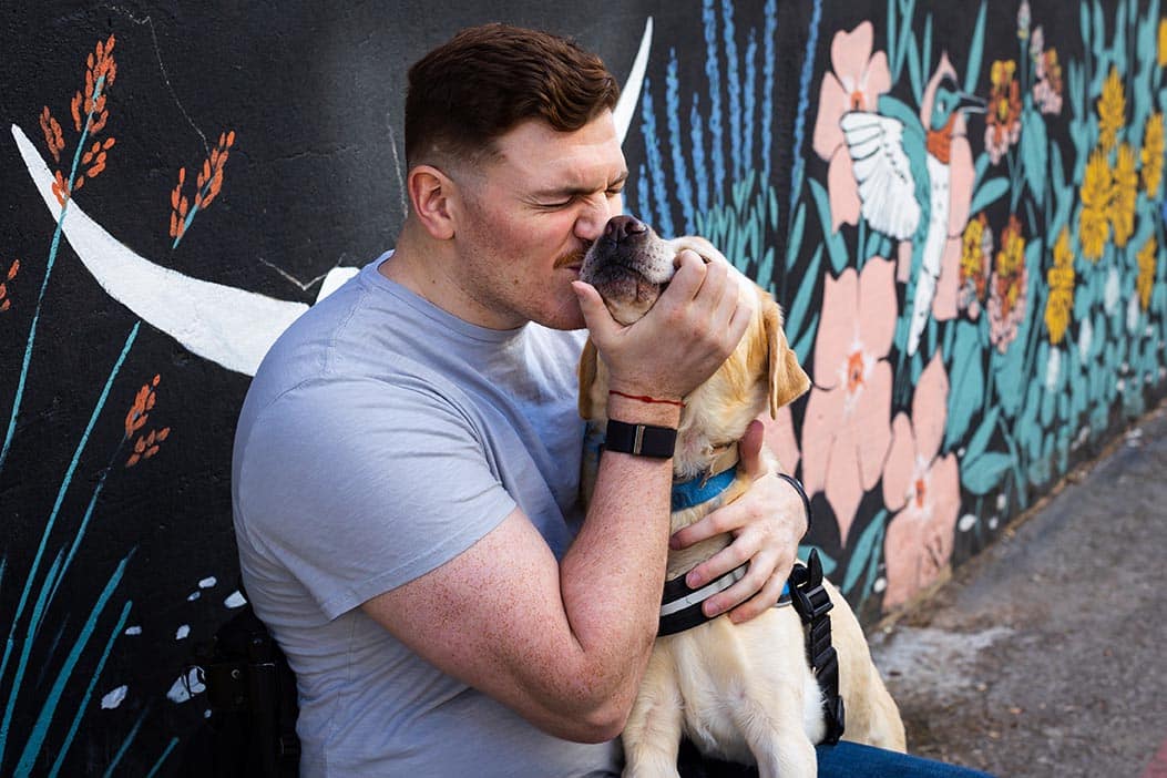 A man hugs and kisses his yellow lab service dog who is wearing a blue vest